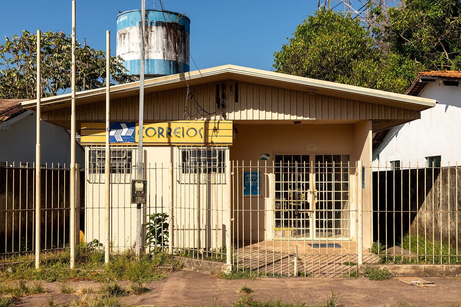 Fachada de uma agência dos Correios em cidade pequena, com letreiro amarelo e portão de grade fechado