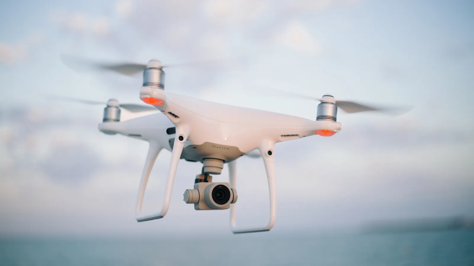 Drone quadricóptero branco com câmera acoplada voando contra um céu com nuvens, com o mar desfocado ao fundo