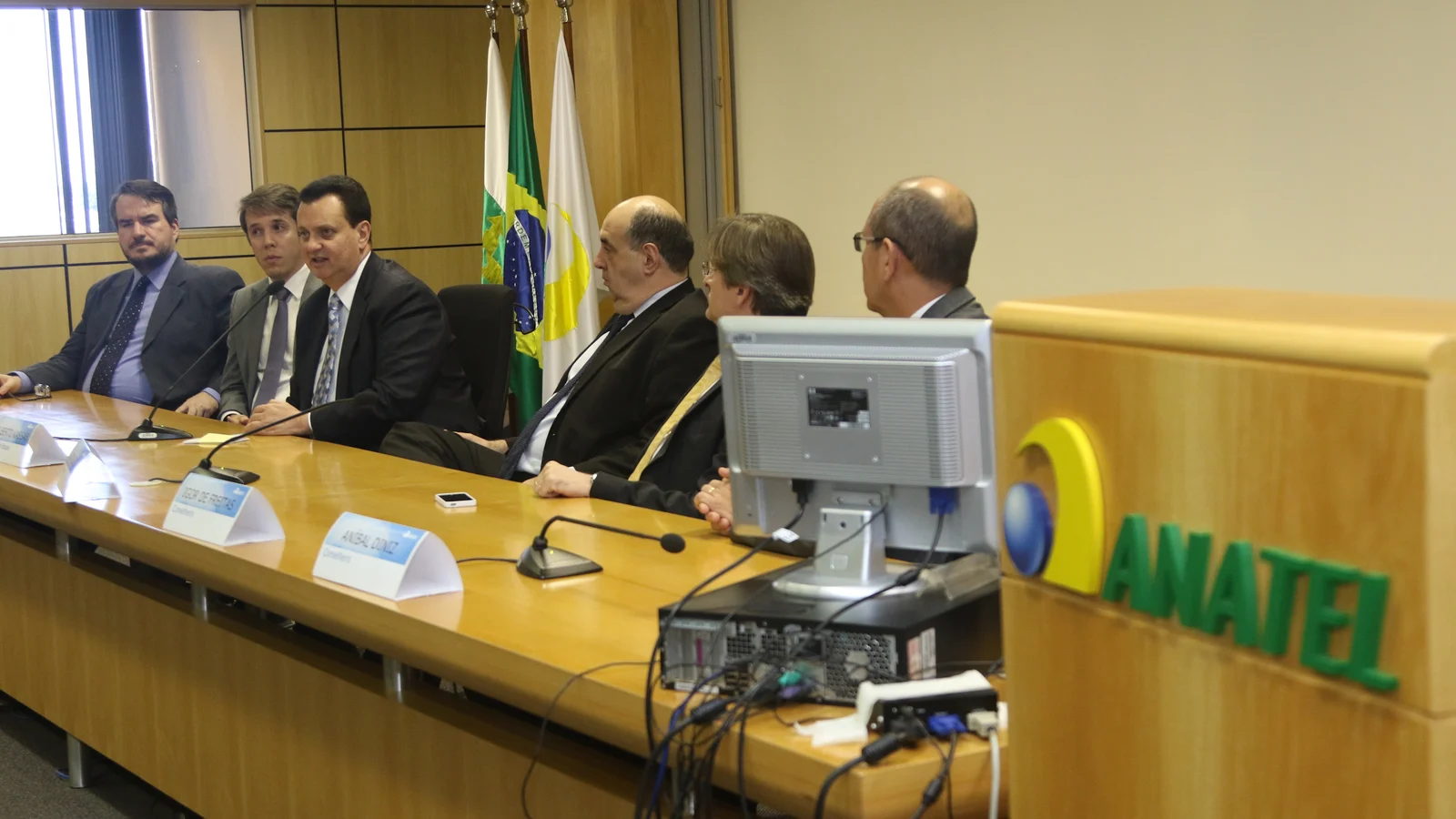 Mesa de reunião com conselheiros da Anatel sentados, bandeiras do Brasil ao fundo e o logotipo ANATEL em destaque à direita