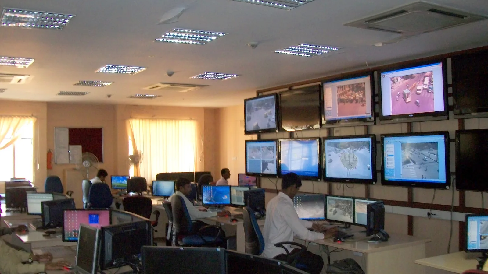 Sala de controle de trânsito com vários operadores em computadores e parede de telas mostrando câmeras de tráfego