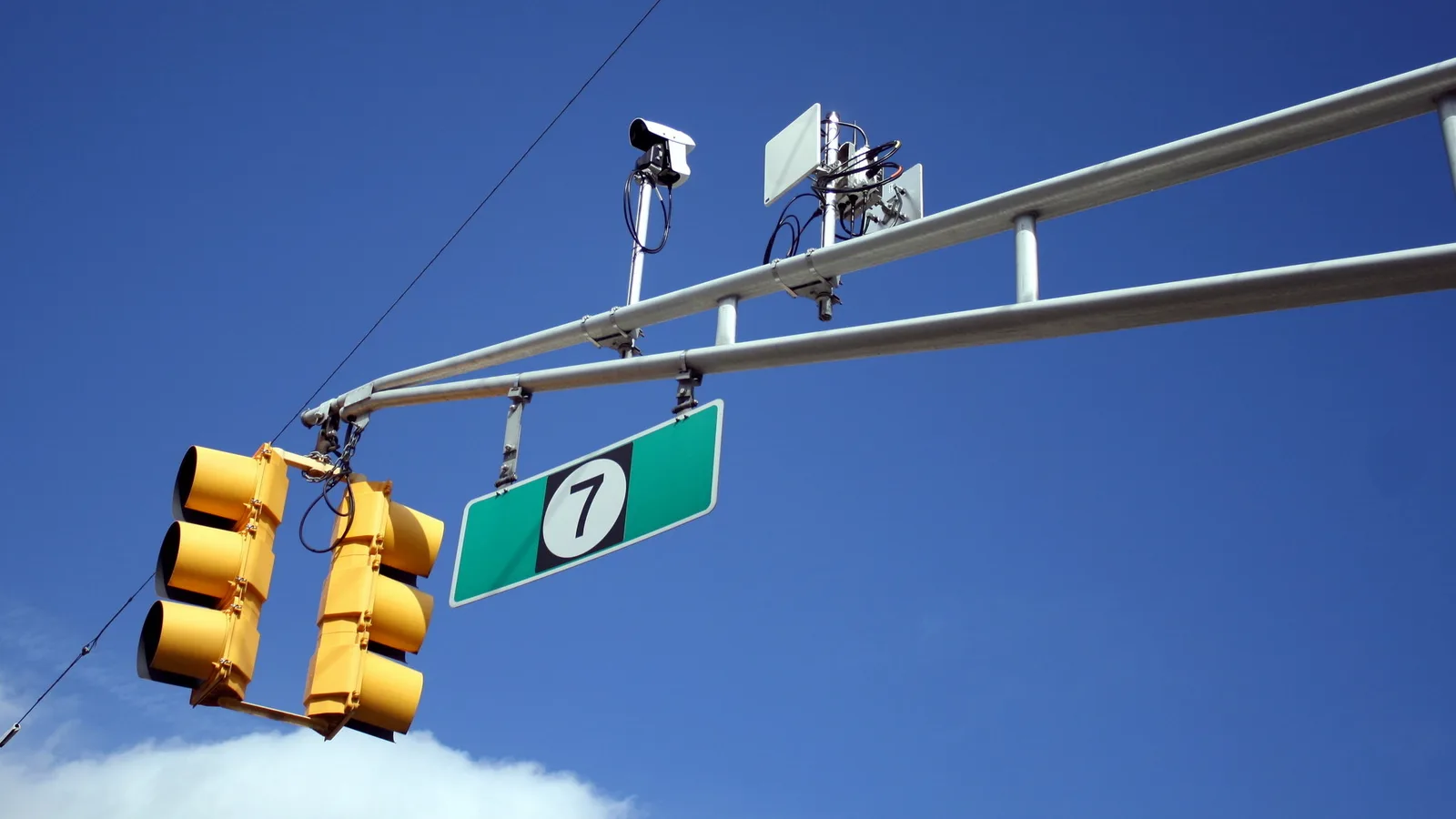 Câmera de fiscalização e antena instaladas em braço de semáforo sobre avenida, com placa de via ao lado