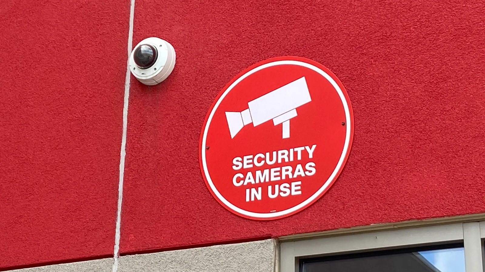 Câmera de segurança tipo dome instalada na fachada de um estabelecimento comercial, ao lado de placa "Security Cameras In Use"