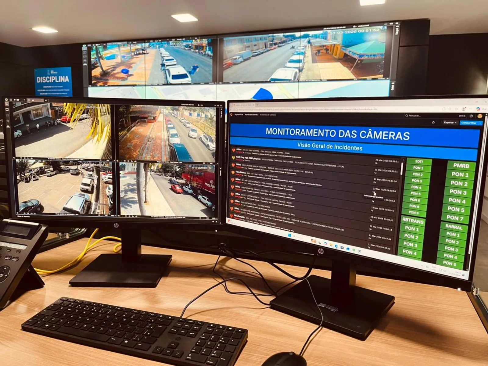 Mesa de trabalho com três monitores mostrando câmeras de segurança ao vivo e um painel chamado Monitoramento das Câmeras, com lista de alertas