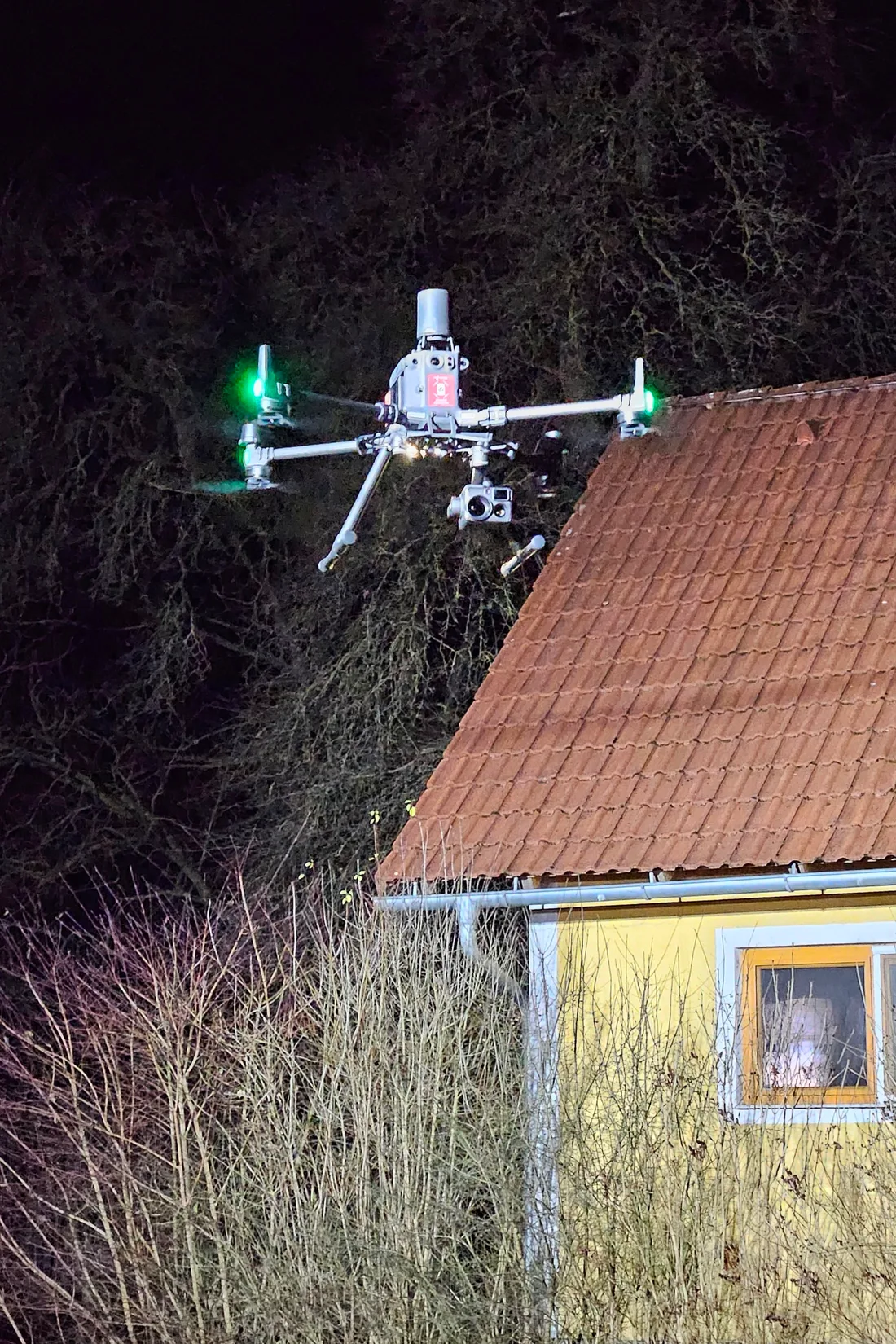 Drone profissional modelo DJI Matrice em voo noturno, com câmera térmica visível, sobre telhado de casa