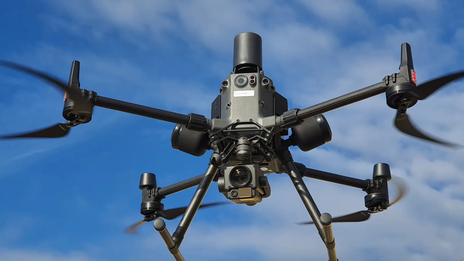 Drone profissional de grande porte em voo contra céu azul, com câmera e sensores visíveis na parte inferior