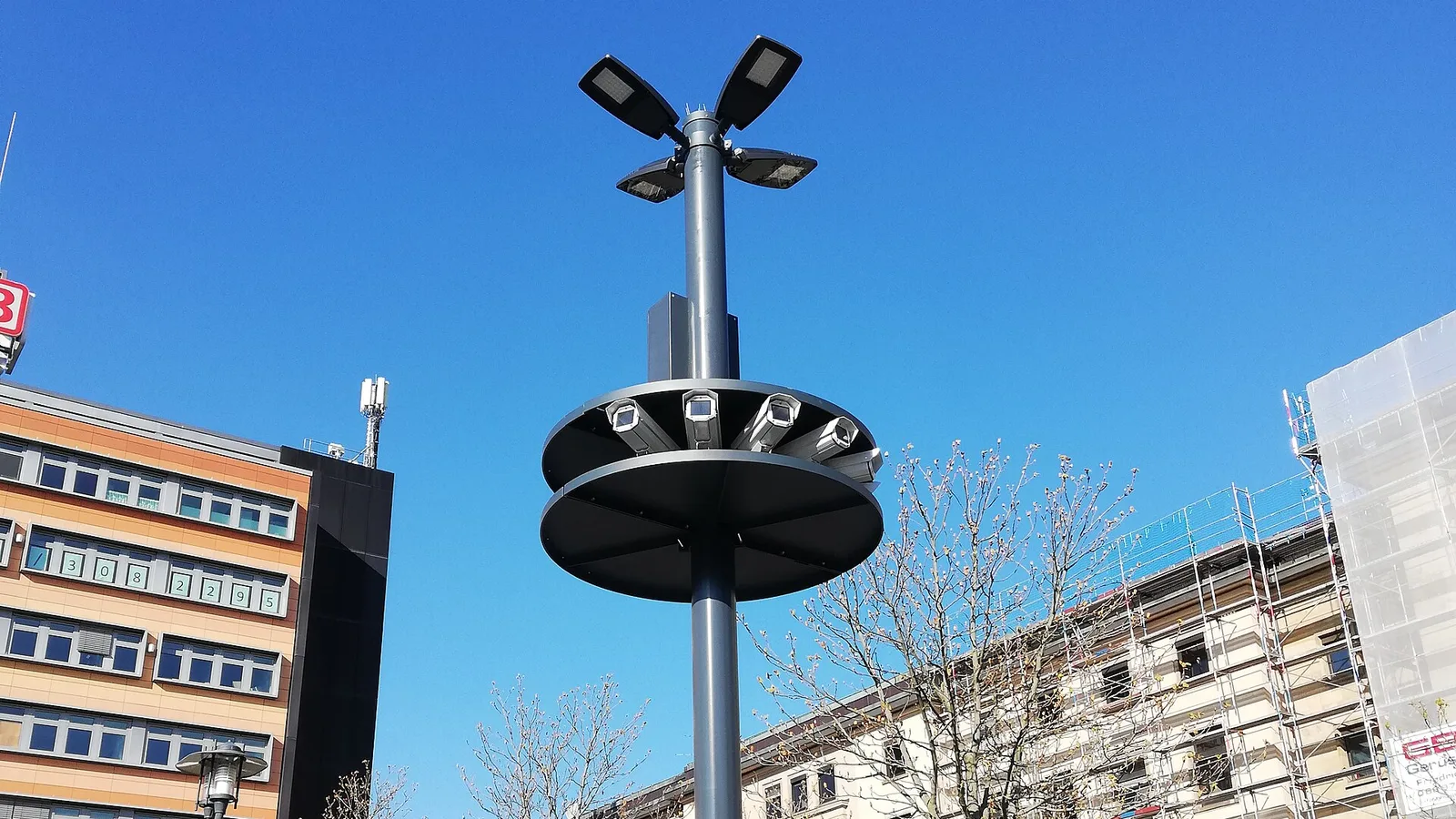 Poste alto em praça pública com um anel metálico contendo várias câmeras de vigilância apontadas para direções diferentes, com prédios ao fundo e céu azul