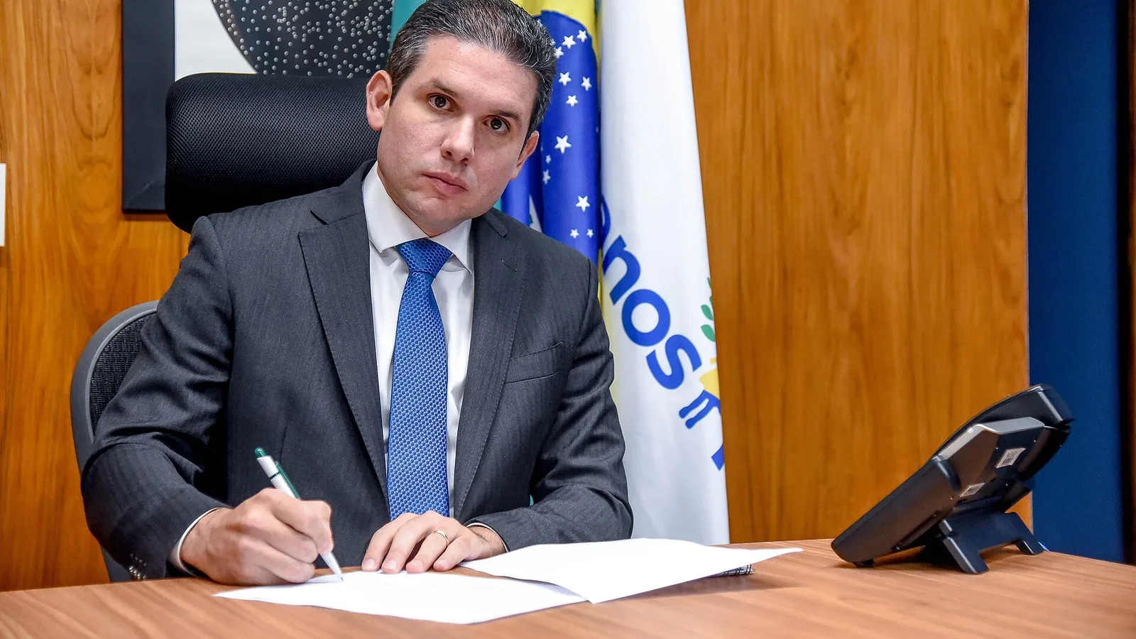 Hugo Motta, presidente da Câmara dos Deputados, sentado a uma mesa de madeira assinando um documento, com bandeiras do Brasil ao fundo