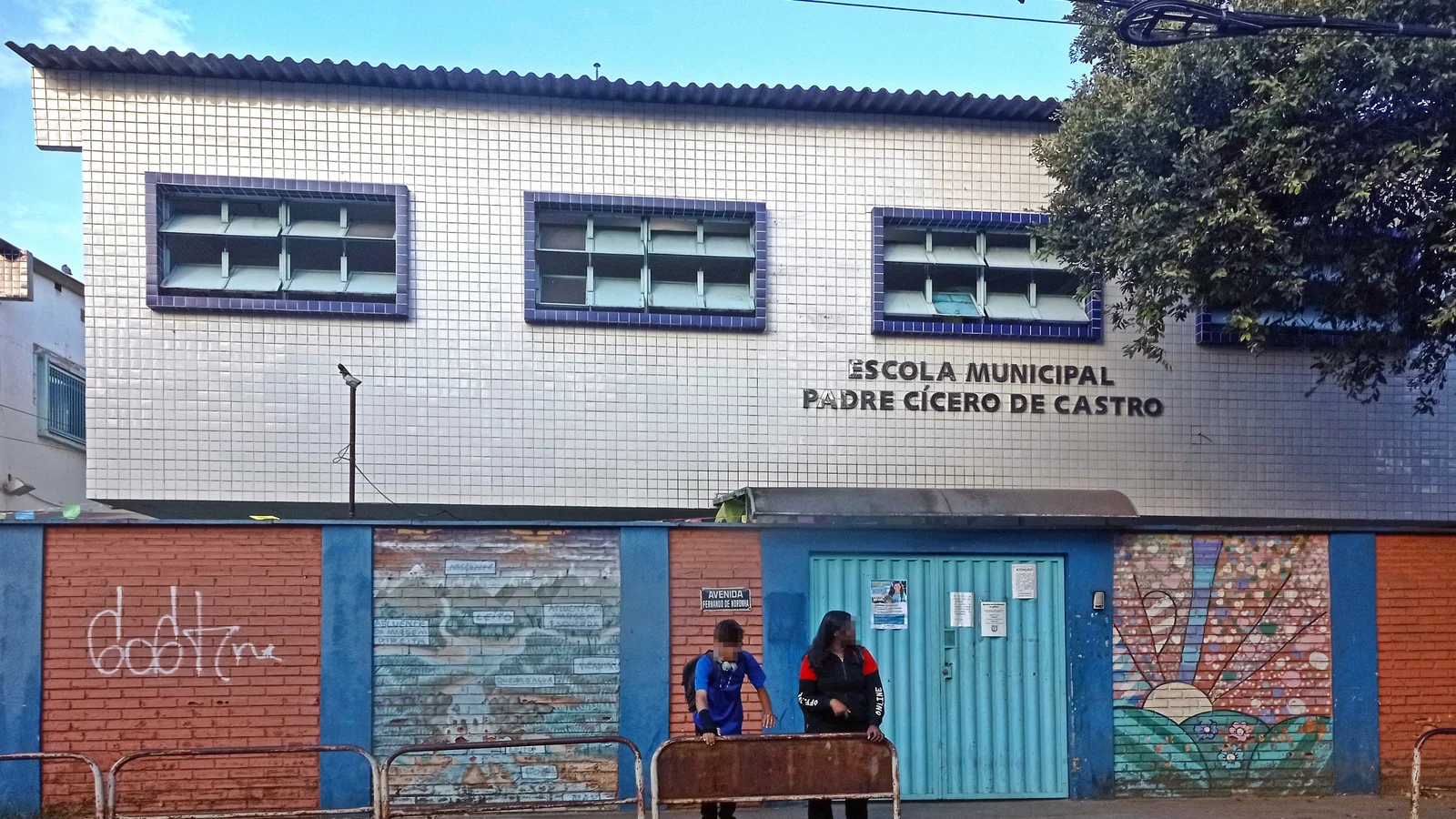 Fachada de escola municipal com pátio e portão de entrada, estudantes na calçada