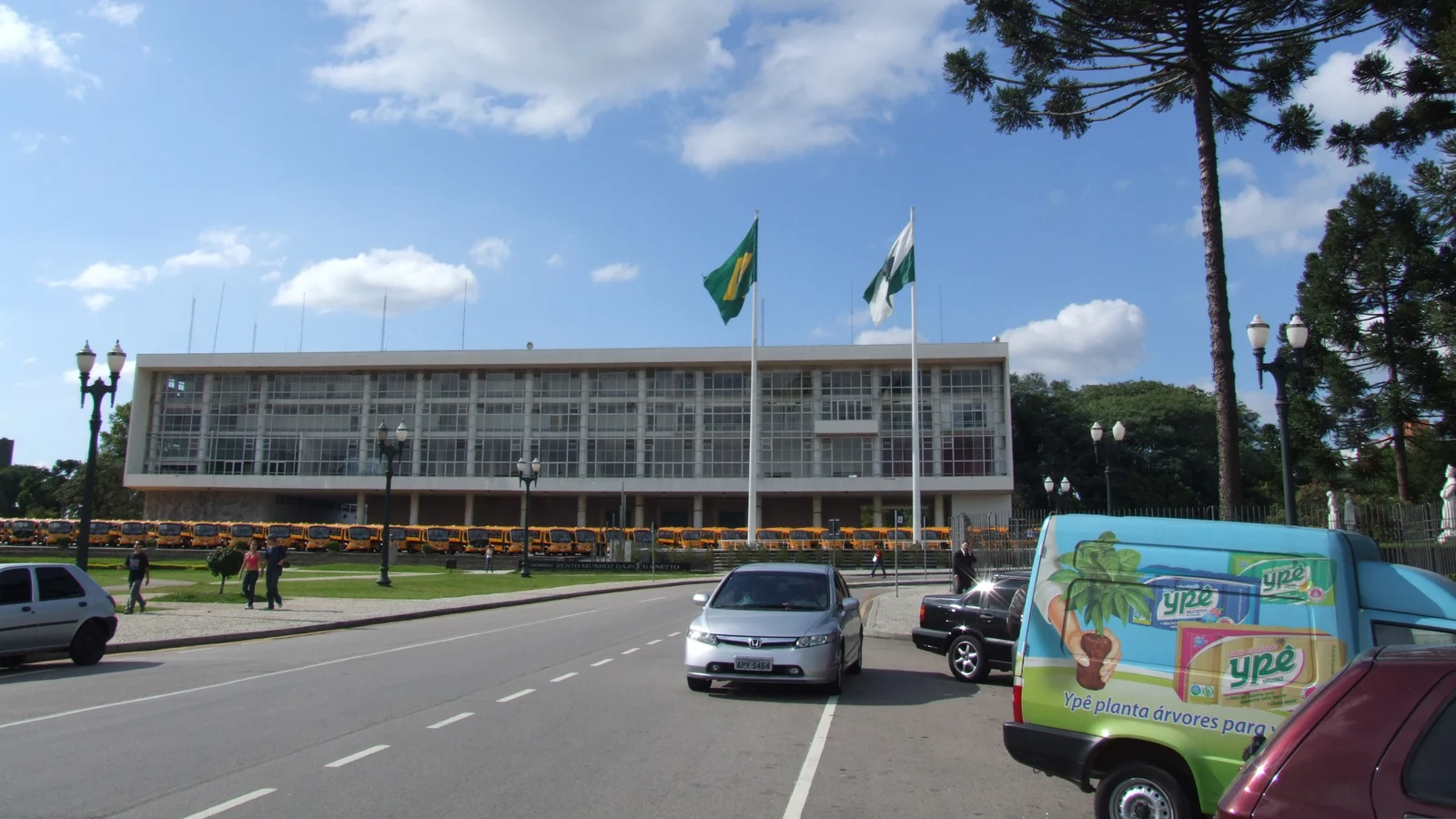Prédio governamental modernista com bandeiras hasteadas e ônibus escolares estacionados em frente