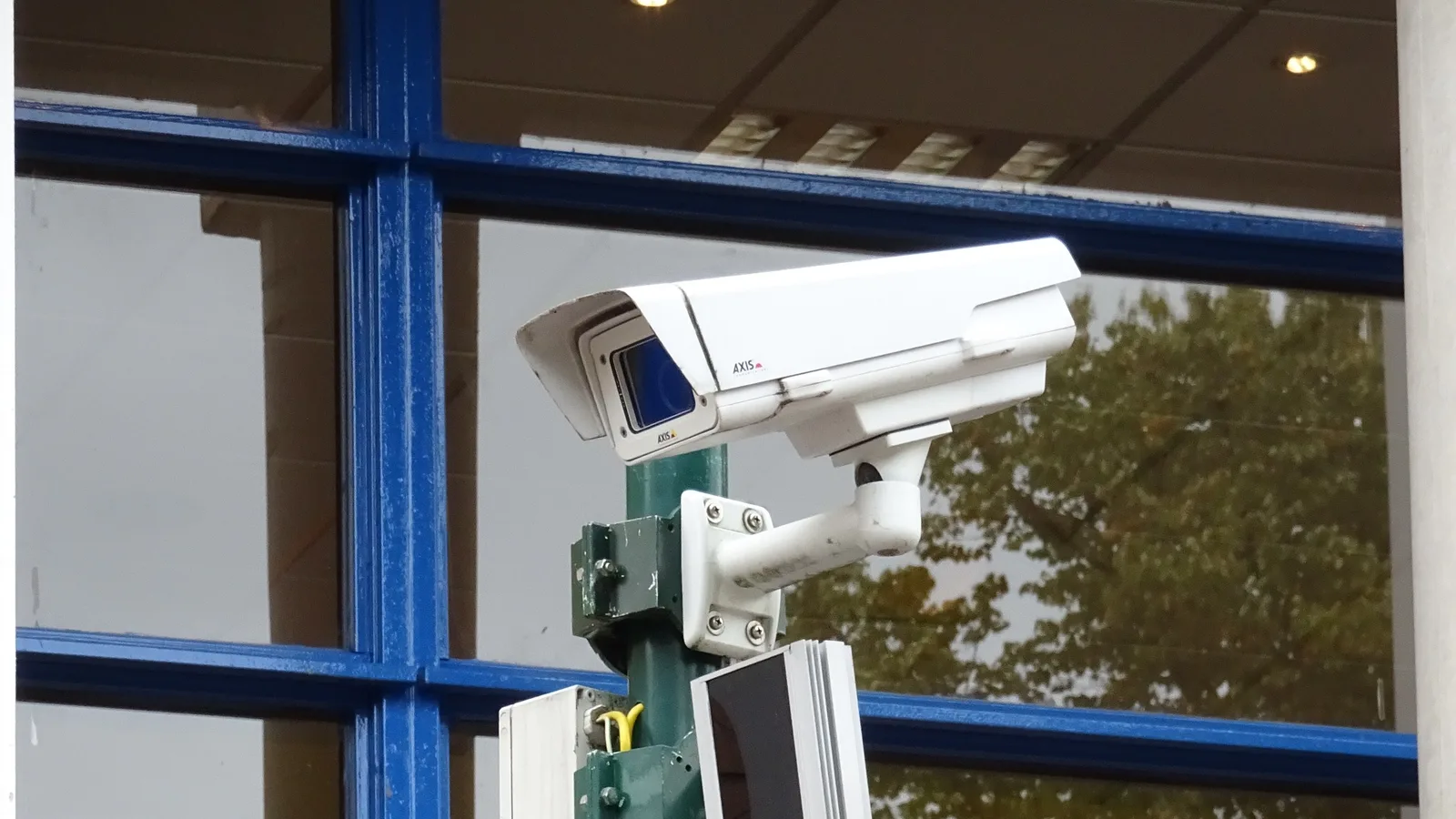 Câmera de vigilância branca instalada em poste, com a marca AXIS visível na carcaça, montada ao lado de um refletor auxiliar contra uma fachada envidraçada