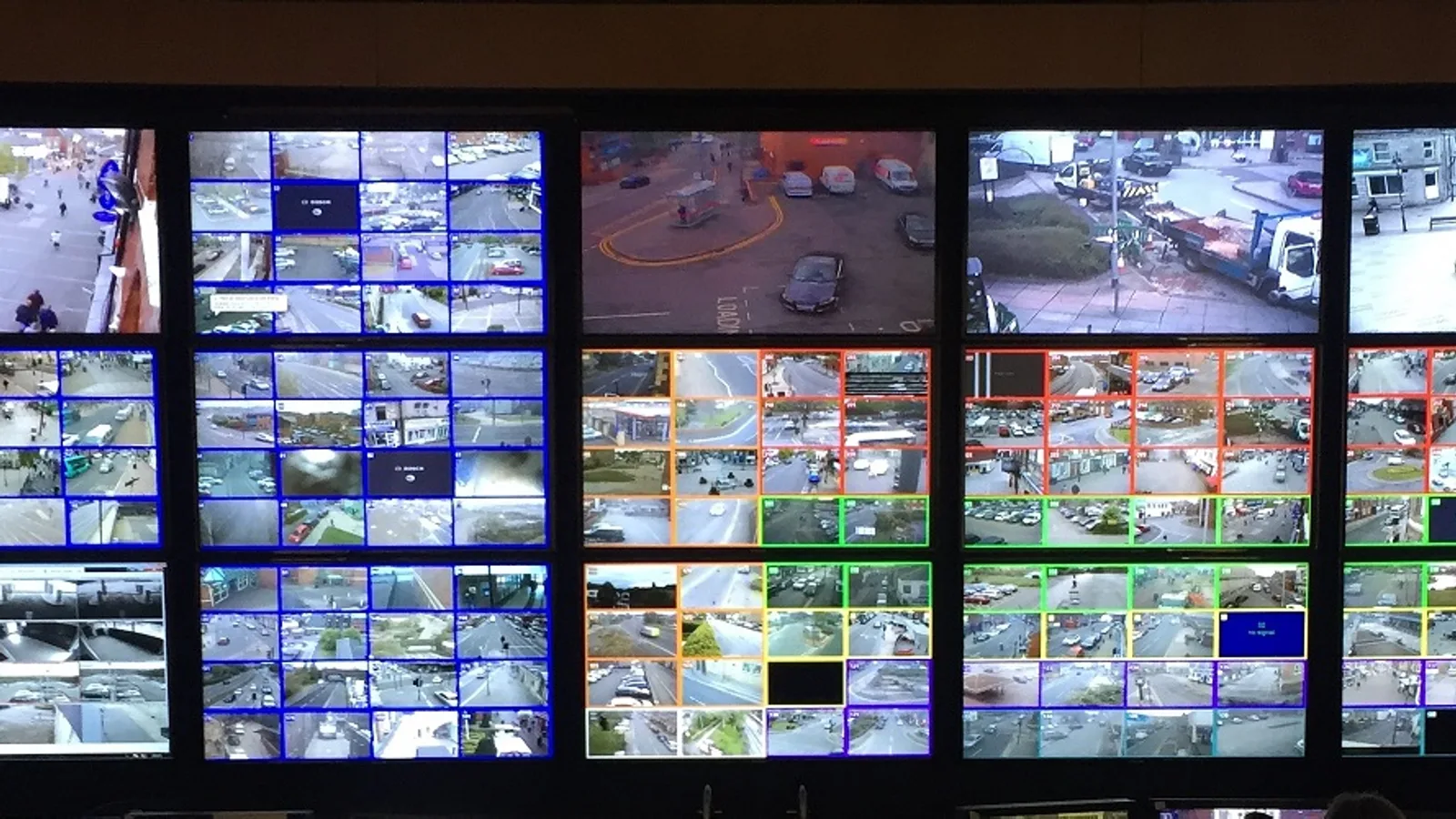 Parede com dezenas de monitores de vigilância exibindo imagens de câmeras de rua, vistas de um centro de controle e monitoramento