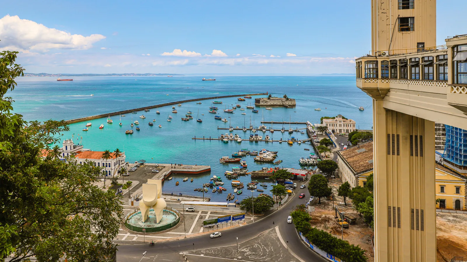 Vista aérea do Elevador Lacerda e da Baía de Todos os Santos em Salvador, com barcos ancorados, o Forte de São Marcelo ao fundo e prédios do Comércio à direita