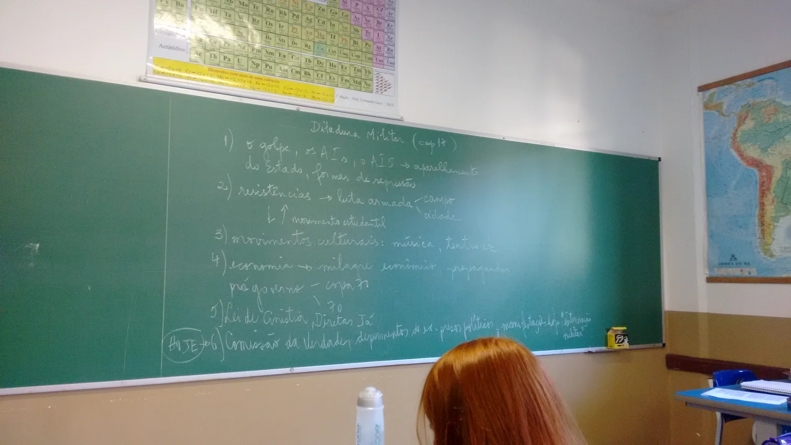 Sala de aula com lousa verde, tabela periódica e mapa do Brasil na parede