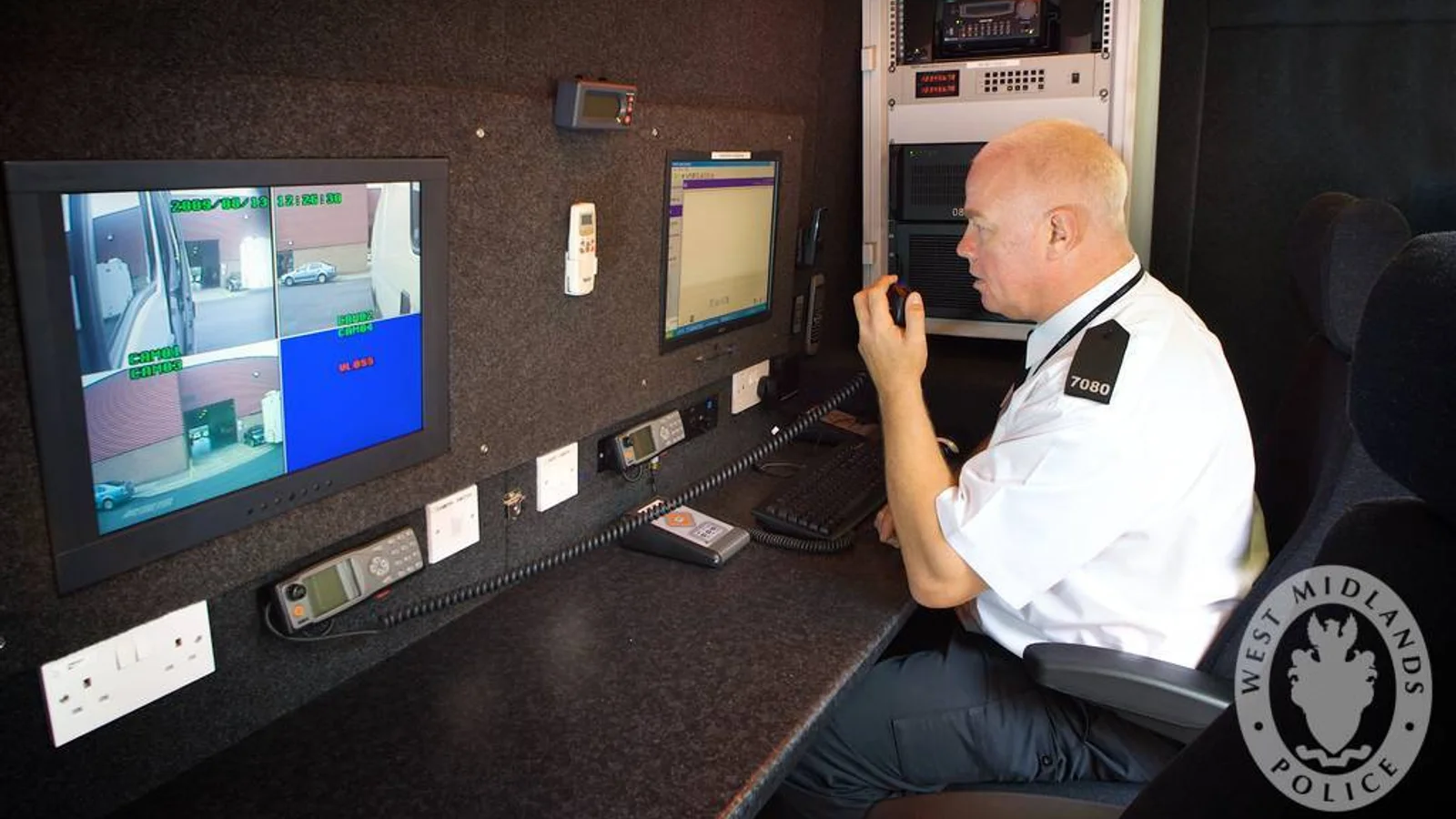 Policial em uma central móvel de monitoramento, com tela dividida em quatro câmeras e rádio na mão