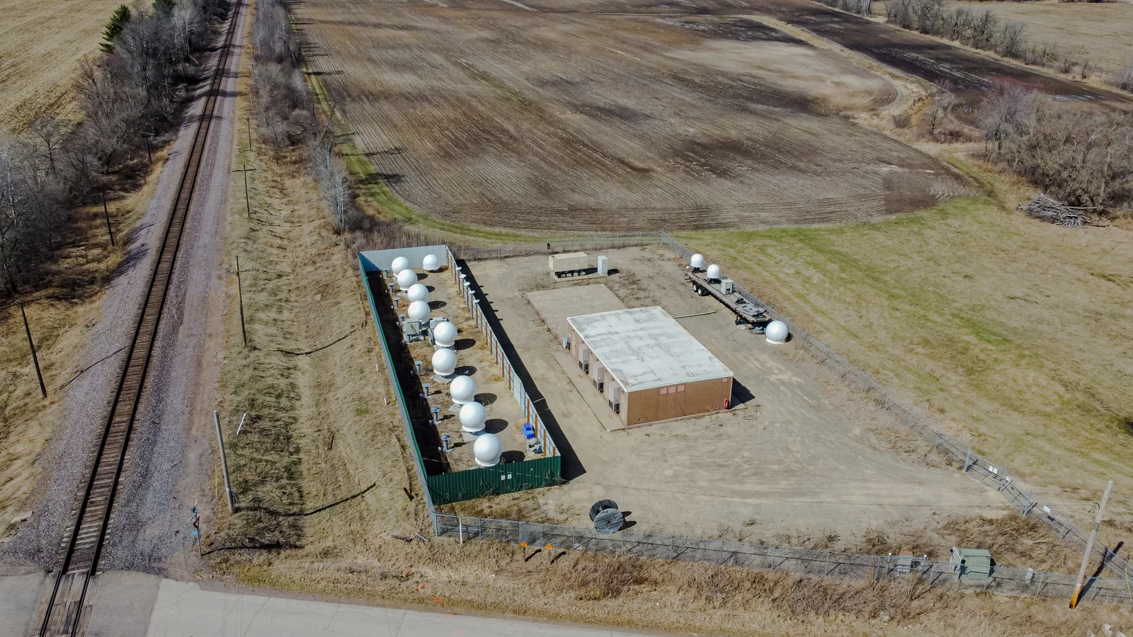 Vista aérea de uma estação terrestre da Starlink, com fileira de antenas parabólicas protegidas por radomes brancos dentro de um perímetro cercado, em meio a campos agrícolas