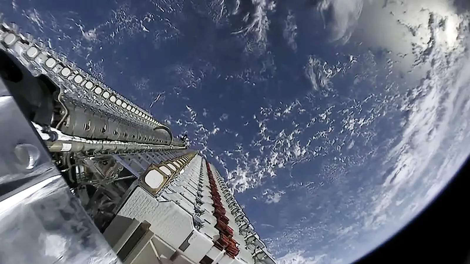 Vista a partir de um foguete Falcon 9 mostrando uma fileira de satélites Starlink empilhados momentos antes de entrar em órbita, com a Terra e nuvens ao fundo