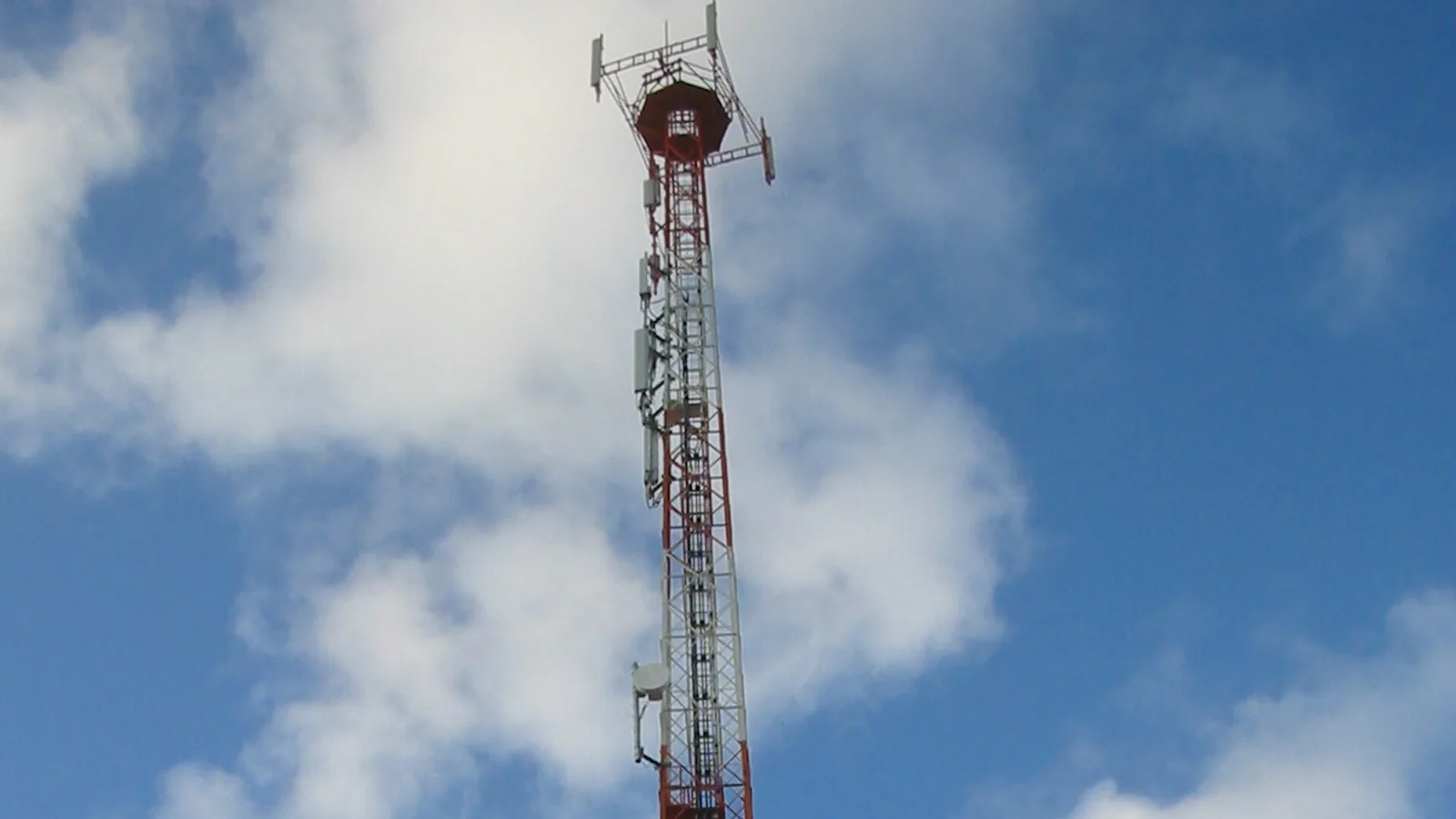 Torre de telefonia celular vermelha e branca contra um céu azul com nuvens, com antenas e equipamentos de transmissão instalados ao longo da estrutura