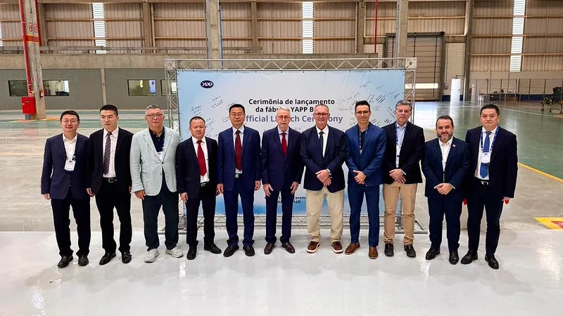 Grupo de cerca de doze pessoas de terno posa para foto diante de um painel branco com o logotipo YAPP, dentro de um galpão industrial de piso claro