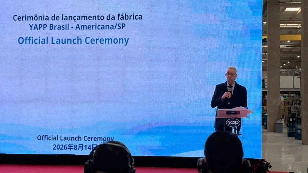 Homem de terno discursa em um púlpito com o logotipo YAPP, diante de um telão que exibe "Cerimônia de lançamento da fábrica YAPP Brasil - Americana/SP", com o interior do galpão industrial à direita