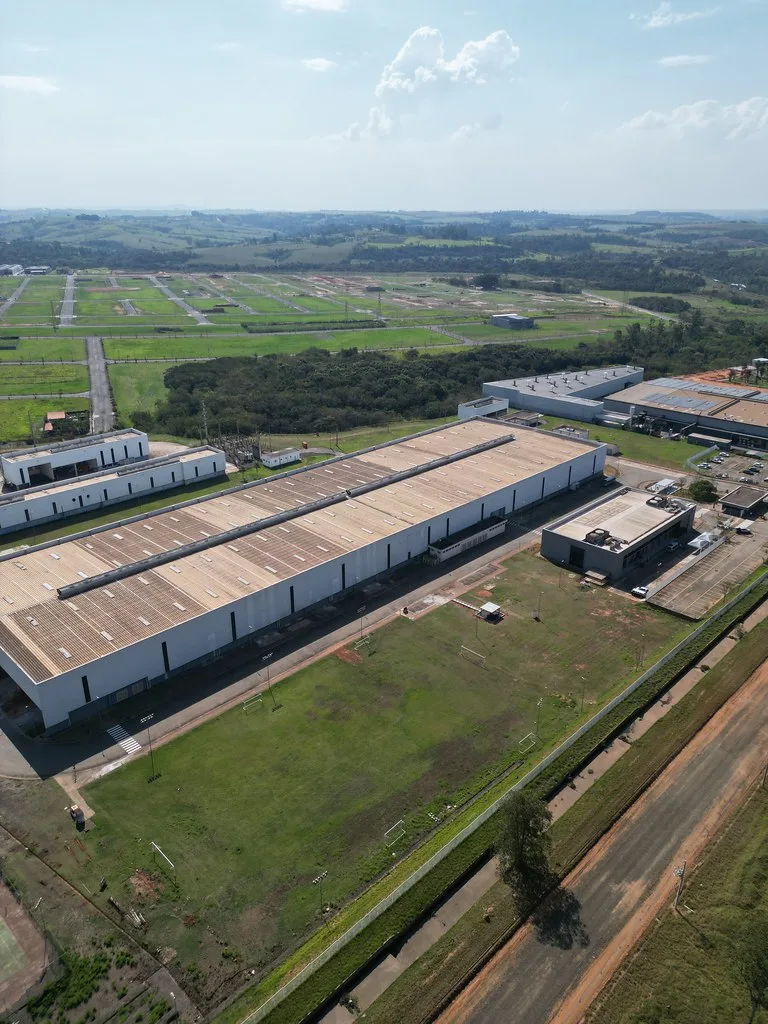 Vista aérea de galpões industriais brancos de telhado longo, cercados por gramado e áreas verdes, com loteamentos e estradas de terra ao fundo