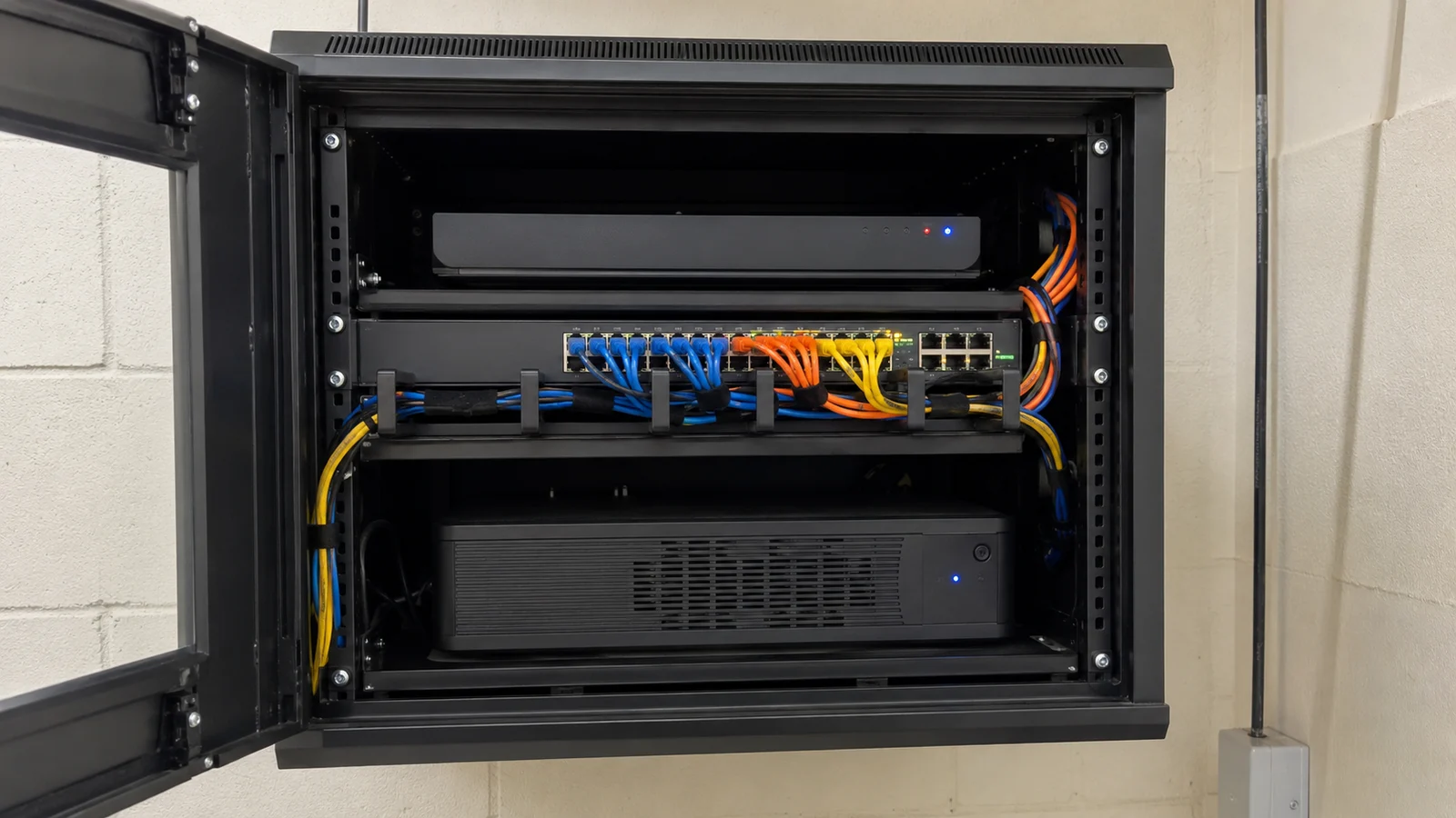 Rack de parede aberto com gravador de imagens no topo, switch de rede com cabos organizados no meio e nobreak na parte inferior
